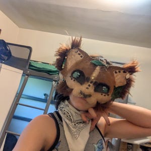 May include: A handmade, brown and tan furry mask with large, expressive eyes and decorative accents. The mask has a cat-like appearance, with pointed ears and a fluffy mane. The mask is adorned with green leaves and small embellishments.