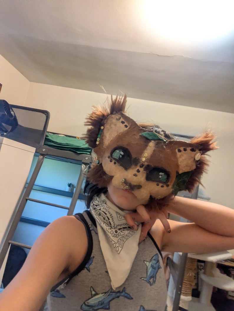 May include: A handmade, brown and tan furry mask with large, expressive eyes and decorative accents. The mask has a cat-like appearance, with pointed ears and a fluffy mane. The mask is adorned with green leaves and small embellishments.
