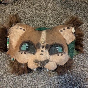 May include: A handmade, brown and tan animal mask with teal eyes, white teeth, and decorative embellishments. The mask has a furry mane and is adorned with beads, leaves, and small metal accents. The mask is designed to be worn.