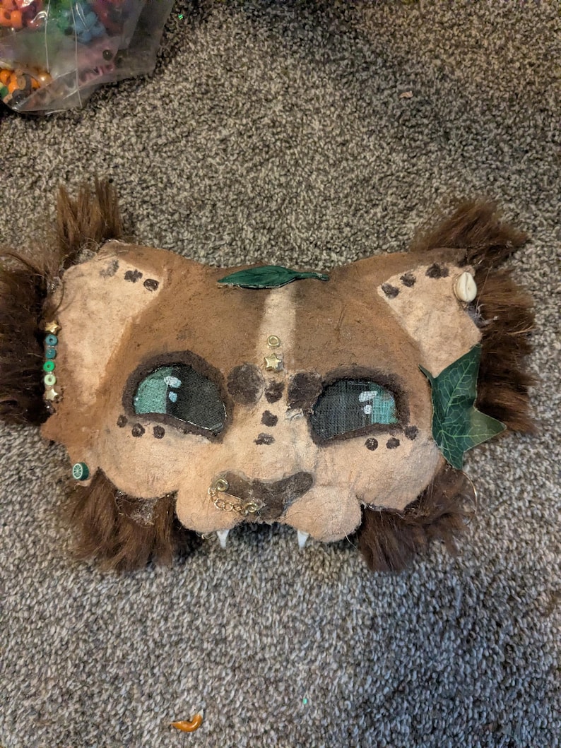 May include: A handmade, brown and tan animal mask with teal eyes, white teeth, and decorative embellishments. The mask has a furry mane and is adorned with beads, leaves, and small metal accents. The mask is designed to be worn.