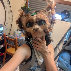 May include: A handmade, brown and tan animal mask with large, teal-colored eyes and a detailed snout. The mask has faux fur accents and is adorned with small decorative elements. The mask is designed to be worn.
