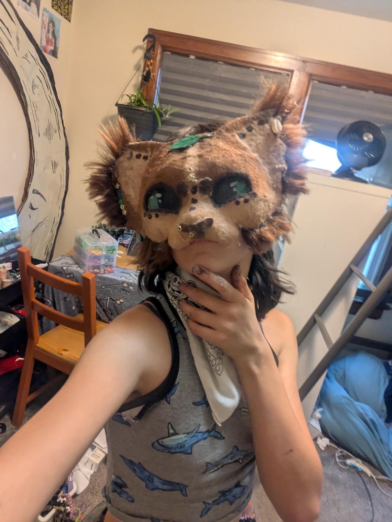 May include: A handmade, brown and tan animal mask with large, teal-colored eyes and a detailed snout. The mask has faux fur accents and is adorned with small decorative elements. The mask is designed to be worn.
