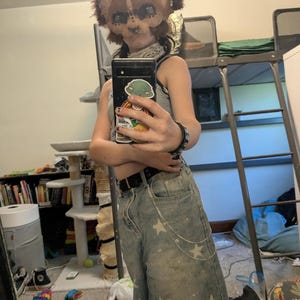 May include: A brown and tan animal mask with large ears and eye details. The person is wearing a gray tank top and light wash denim shorts with white star patterns and a silver chain. A black belt and bracelet are also visible.