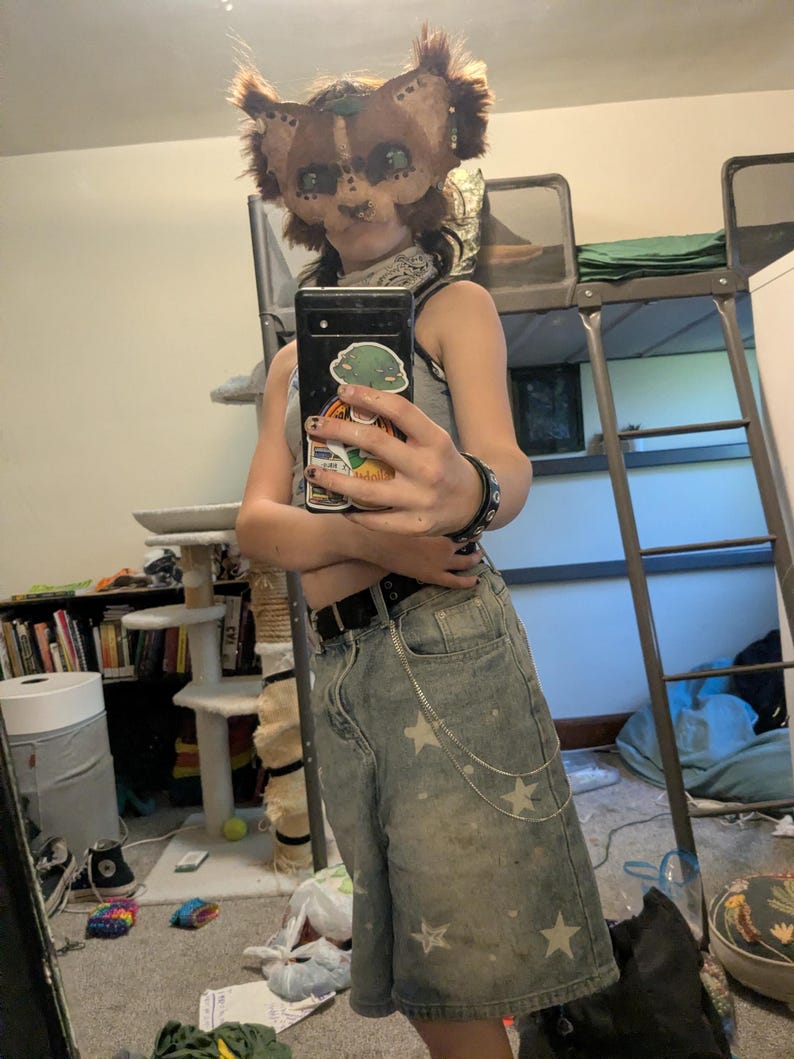 May include: A brown and tan animal mask with large ears and eye details. The person is wearing a gray tank top and light wash denim shorts with white star patterns and a silver chain. A black belt and bracelet are also visible.