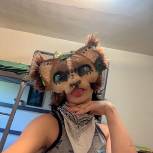 May include: A handmade, brown and tan feline mask with green eyes, a nose ring, and faux fur accents. The mask features decorative leaves and is worn with a white bandana and a gray tank top with a shark pattern.