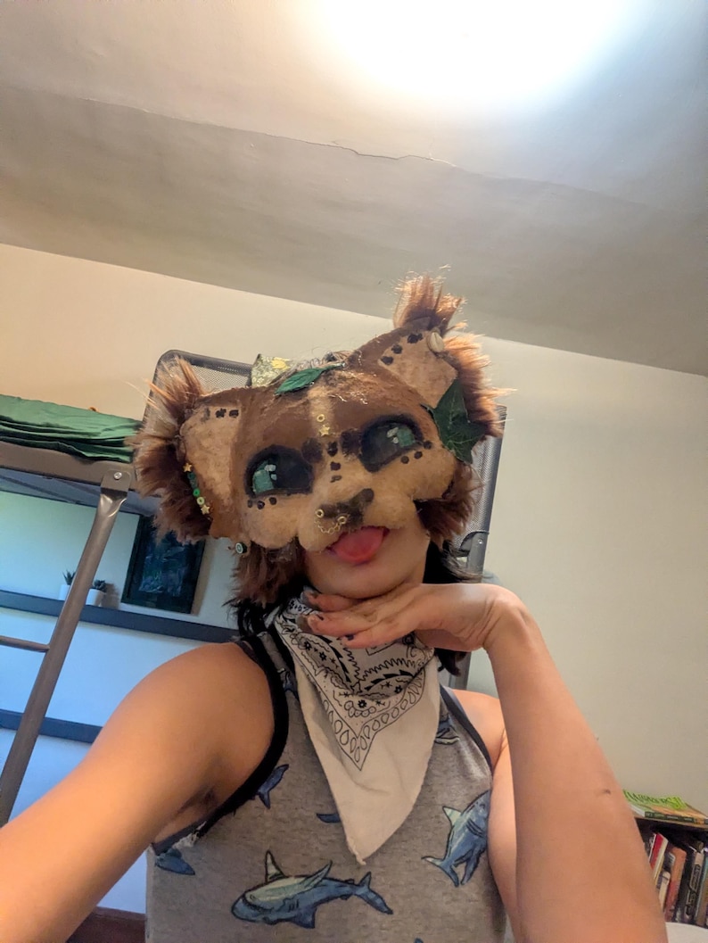 May include: A handmade, brown and tan feline mask with green eyes, a nose ring, and faux fur accents. The mask features decorative leaves and is worn with a white bandana and a gray tank top with a shark pattern.