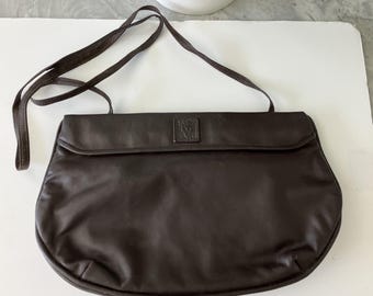 May include: A dark brown leather crossbody bag with a long, thin strap. The bag has a rounded bottom and a flap closure. A small, square emblem is centered on the flap. The bag is made of a smooth, shiny leather.