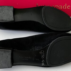 May include: A pair of black suede Kate Spade flats, viewed from the soles. The shoes have rounded toes and rubber soles. The brand name is visible on the soles and the box in the background.