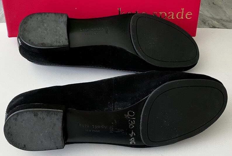 May include: A pair of black suede Kate Spade flats, viewed from the soles. The shoes have rounded toes and rubber soles. The brand name is visible on the soles and the box in the background.
