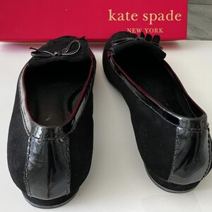 May include: Pair of black suede flats with patent leather accents and small bows. The shoes have a red trim and are from Kate Spade New York.