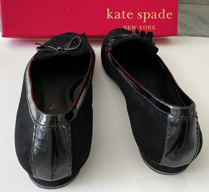 May include: Pair of black suede flats with patent leather accents and small bows. The shoes have a red trim and are from Kate Spade New York.