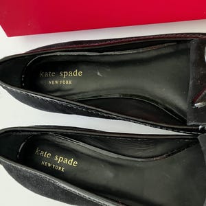 May include: Close-up of two black suede loafers with a glossy black trim. The interior of each shoe is black with gold lettering that reads "kate spade NEW YORK". A small bow detail is visible on the top of one shoe.