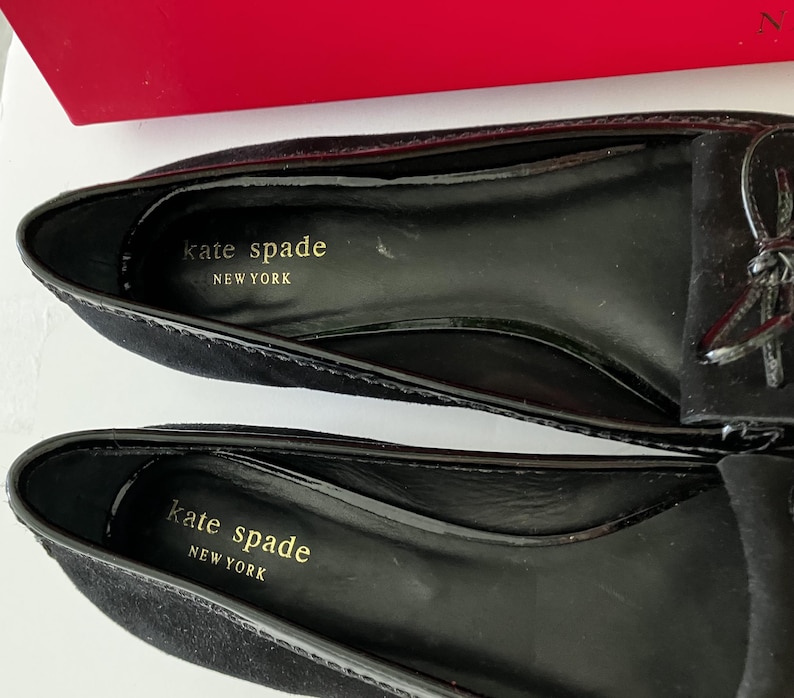 May include: Close-up of two black suede loafers with a glossy black trim. The interior of each shoe is black with gold lettering that reads "kate spade NEW YORK". A small bow detail is visible on the top of one shoe.