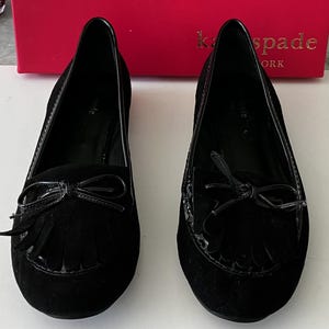 May include: A pair of black suede flats with a bow and fringe detail. The shoes have a glossy trim and are displayed in front of a red box with gold lettering.