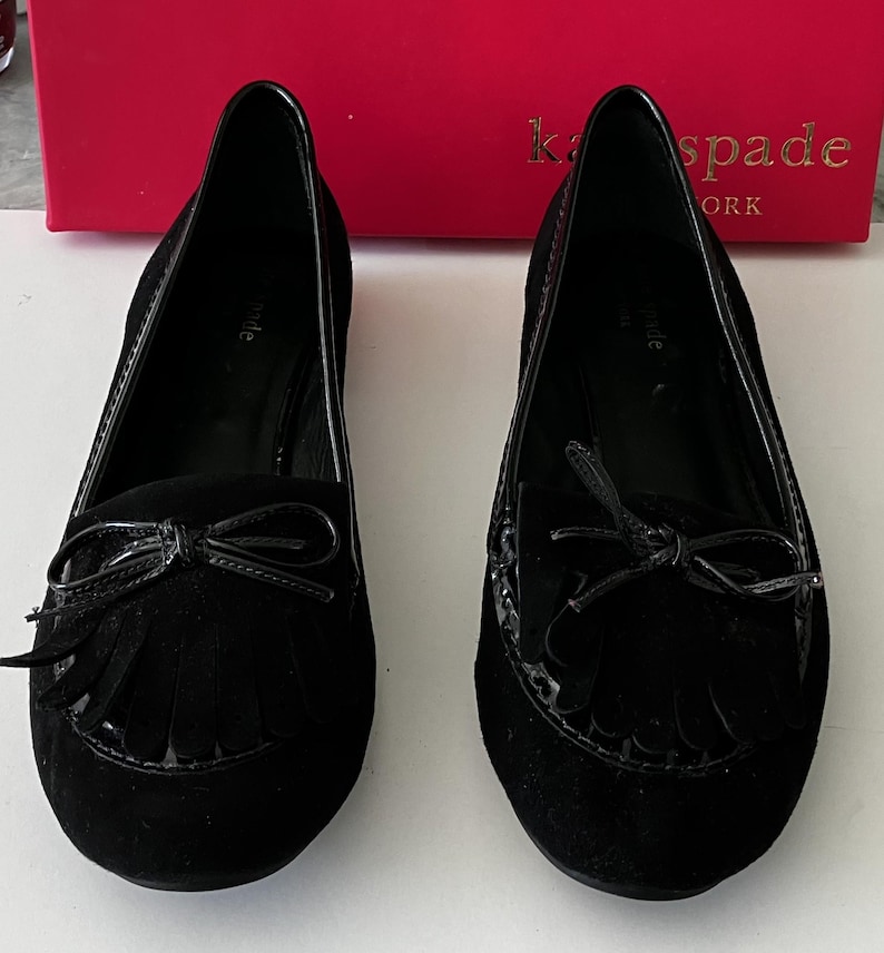 May include: A pair of black suede flats with a bow and fringe detail. The shoes have a glossy trim and are displayed in front of a red box with gold lettering.