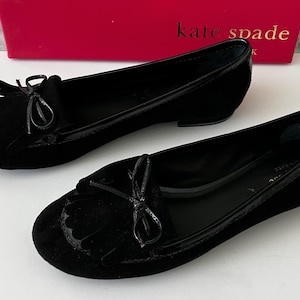 May include: A pair of black suede loafers with a glossy patent leather trim. Each shoe features a decorative bow and fringe detail on the vamp. The shoes are set against a white surface, with a red box bearing the "kate spade" logo in the background.