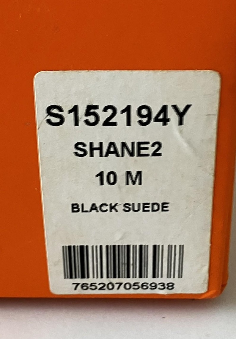May include: A white label with black text against an orange background. The label reads "S152194Y SHANE2 10 M BLACK SUEDE" and includes a barcode and the number "765207056938".