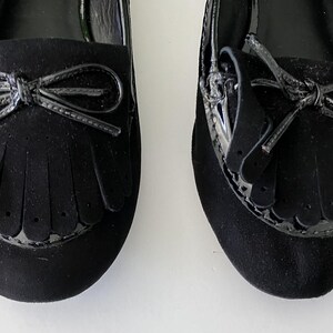 May include: Pair of black suede loafers with patent leather accents. Each shoe features a bow detail and fringe across the vamp. The shoes have a classic, elegant design, suitable for various occasions.