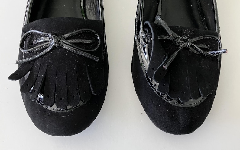 May include: Pair of black suede loafers with patent leather accents. Each shoe features a bow detail and fringe across the vamp. The shoes have a classic, elegant design, suitable for various occasions.