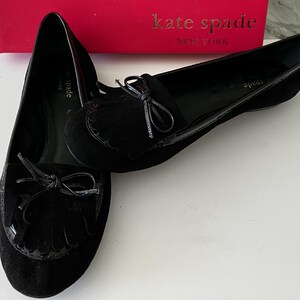 May include: A pair of black suede Kate Spade loafers with bow accents and fringe detailing. The shoes have a glossy trim and are displayed on a white surface, with a red Kate Spade box in the background.