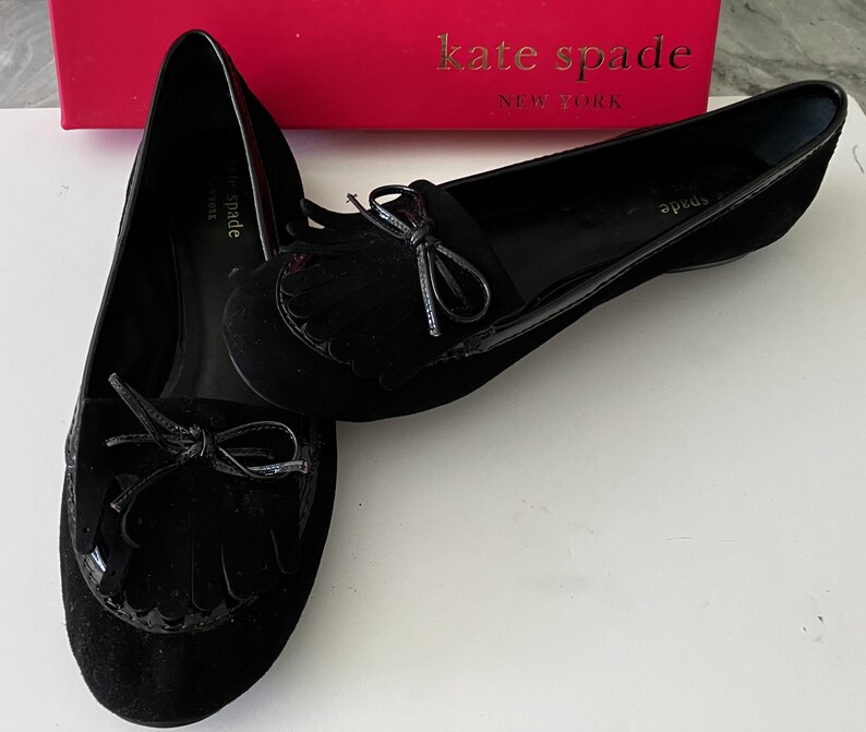 May include: A pair of black suede Kate Spade loafers with bow accents and fringe detailing. The shoes have a glossy trim and are displayed on a white surface, with a red Kate Spade box in the background.