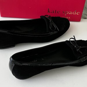 May include: A pair of black suede flats with a glossy patent leather trim. Each shoe features a small bow detail on the vamp. The shoes are displayed in front of a red Kate Spade New York box.