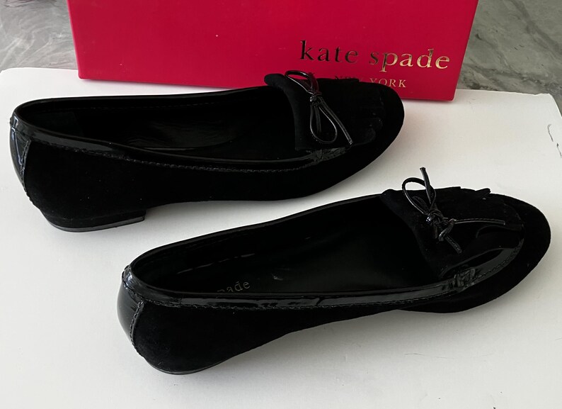 May include: A pair of black suede flats with a glossy patent leather trim. Each shoe features a small bow detail on the vamp. The shoes are displayed in front of a red Kate Spade New York box.