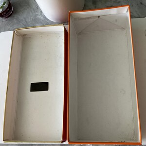 May include: Open rectangular box with a white interior and gold trim. The exterior is orange with a white interior. A small black rectangle is inside the box. The box is open and empty.
