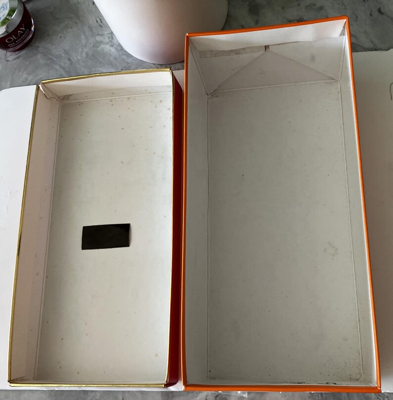 May include: Open rectangular box with a white interior and gold trim. The exterior is orange with a white interior. A small black rectangle is inside the box. The box is open and empty.