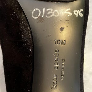 May include: Black shoe sole with the brand name "kate spade NEW YORK" embossed. The size is 10M. Handwritten numbers "0130 596" are visible. The shoe appears to be made of a soft, suede-like material.