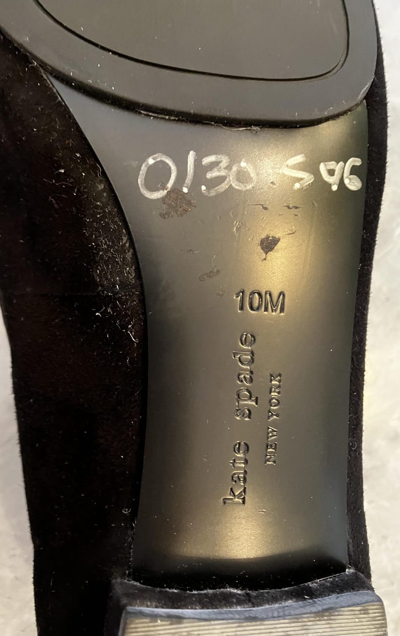 May include: Black shoe sole with the brand name "kate spade NEW YORK" embossed. The size is 10M. Handwritten numbers "0130 596" are visible. The shoe appears to be made of a soft, suede-like material.