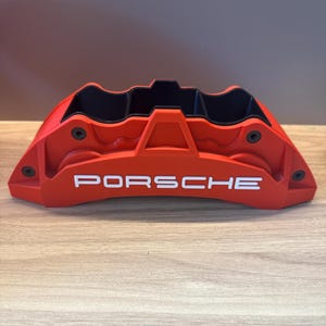 Op de afbeelding: Een felrode Porsche-remklauw met een opvallend wit Porsche-logo in reliëf op de voorzijde. Het onderdeel heeft een matte afwerking met zwarte details aan de binnenzijde en vier zichtbare ronde bevestigingspunten op het buitenste frame.