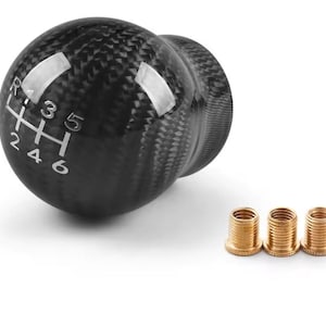 May include: A spherical gear shift knob with a black carbon fiber pattern and a white six-speed manual transmission shift pattern diagram printed on the top. Three small, threaded brass-colored metal adapters are positioned next to the knob.