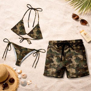 Op de afbeelding: Een camouflage bikini en zwembroek set liggen op zand. De bikini heeft een driehoekige top en een broekje met strikjes. De zwembroek heeft een tailleband met trekkoord. Zonnebril, zonnebrandcrème, een hoed en schelpen zijn ook aanwezig.