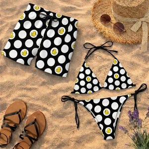 May include: A black and white polka dot bikini and swim trunks set with yellow duck graphics. The image also includes a straw hat, sunglasses, and brown sandals, all arranged on a sandy surface.