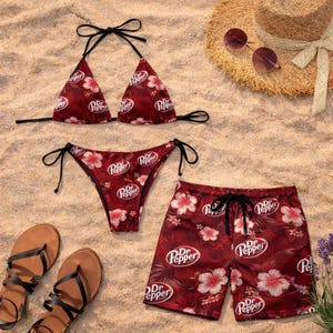May include: A red bikini top and bottom, and matching swim trunks, all featuring a floral pattern and the Dr Pepper logo. The items are arranged on a sandy surface with sandals, a straw hat, and sunglasses.