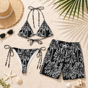 Op de afbeelding: Een zwart-witte bikini set met palmboomprint en bijpassende zwembroek worden gepresenteerd op een zanderige ondergrond. De bikinitop heeft halterbandjes en de broekjes hebben strikzijden. De zwembroek heeft een taille met trekkoord.