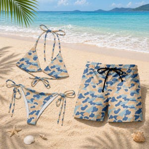 Può includere: Un set di costumi da bagno a tre pezzi con un motivo mimetico nei toni del blu, beige e crema. Il set comprende un top bikini, uno slip bikini e un pantaloncino da bagno con coulisse. Gli articoli sono esposti su una spiaggia sabbiosa.