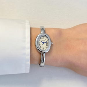 May include: Silver oval wristwatch with a cream-colored face and blue hands. The watch features Roman numerals and the brand name "NAIDU". The watch is worn as a bracelet. The watch is on a wrist.