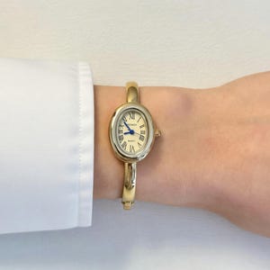 May include: Gold-toned oval wristwatch with a cream-colored face and blue hands. The watch features Roman numeral hour markers and a gold bangle-style band. The watch is worn on a wrist.