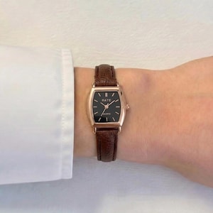 Puede incluir: Un reloj con correa de cuero marrón, caja rectangular dorada y esfera negra. El reloj tiene la palabra "RATE" impresa en la esfera. El reloj se lleva en la muñeca.