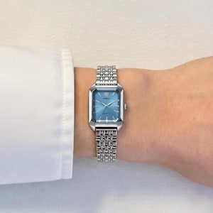 May include: A silver wristwatch with a rectangular face and a light blue dial. The watch has a linked metal band and the brand name "XIMILI" is visible on the watch face. The watch is worn on a wrist.