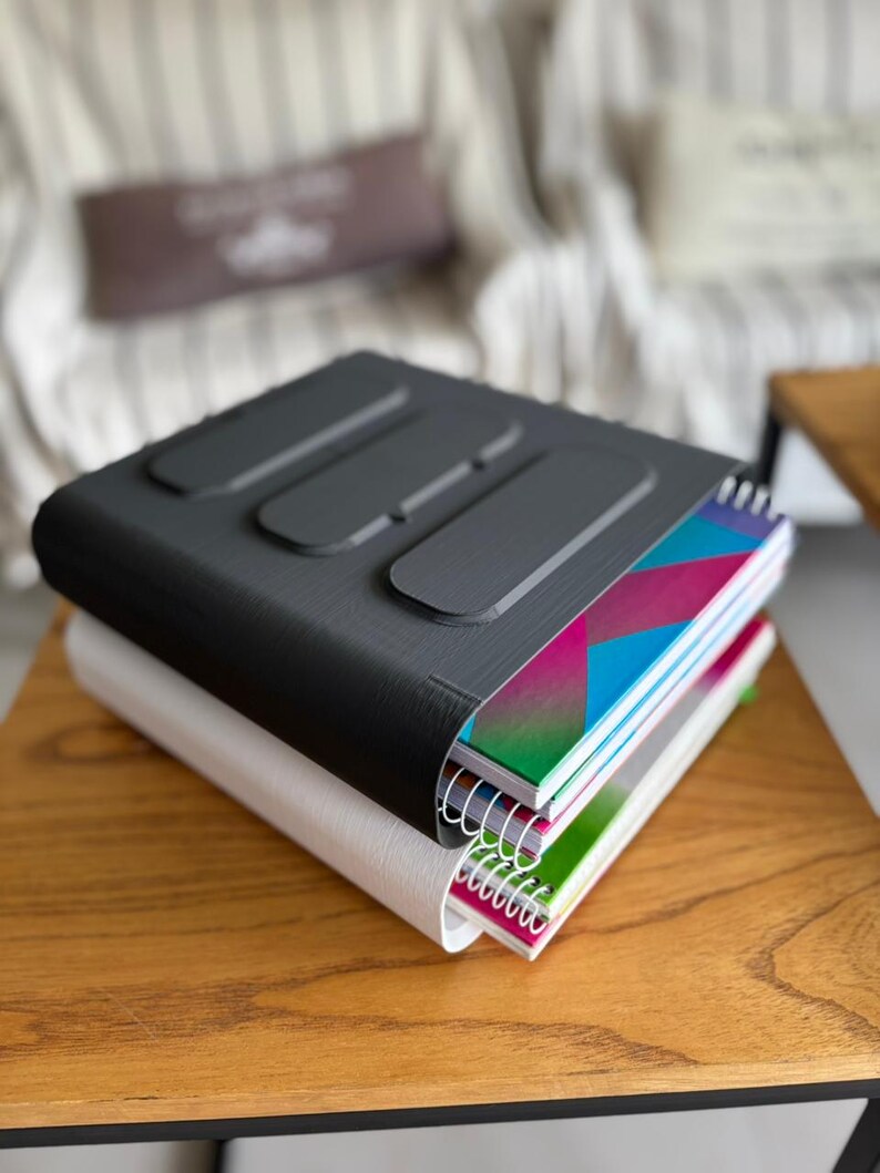 May include: A black and white plastic binder set featuring a textured exterior with three raised rectangular accents on the top cover. The binders are stacked together and contain spiral-bound notebooks with colorful geometric patterns on the covers.