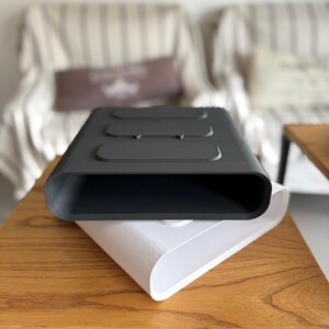 May include: A matte black rectangular container with rounded edges and three raised oval sections on the top surface, stacked directly on top of an identical white container. Both objects feature a hollow, open-ended design with smooth surfaces.