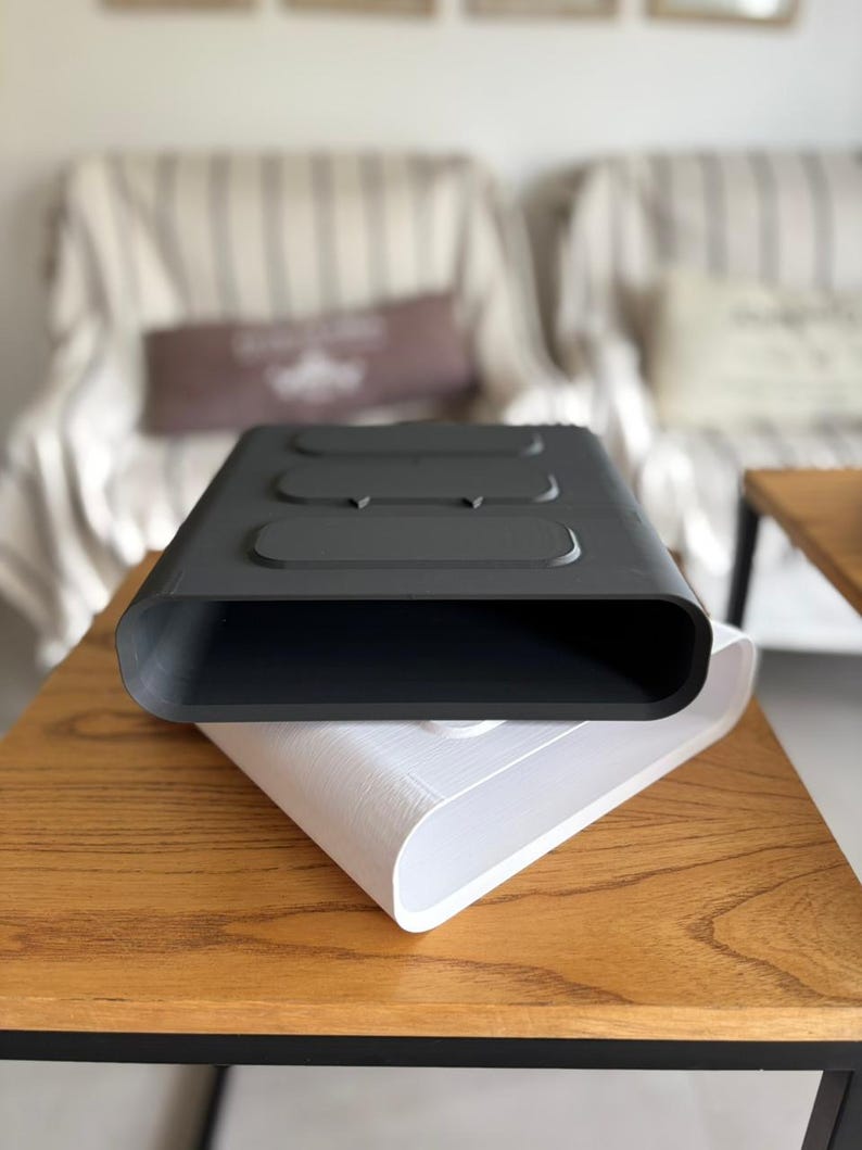 May include: A matte black rectangular container with rounded edges and three raised oval sections on the top surface, stacked directly on top of an identical white container. Both objects feature a hollow, open-ended design with smooth surfaces.
