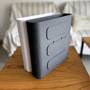 May include: A white and a dark gray vertical storage organizer with rounded edges. The dark gray unit features three horizontal, recessed oval panels on its front face, while the white unit is positioned directly behind it.