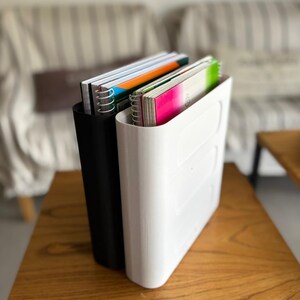 May include: Two vertical document organizers, one matte black and one glossy white, each featuring a smooth, rounded rectangular design. These organizers are filled with spiral-bound notebooks that have colorful covers in shades of orange, green, and pink.