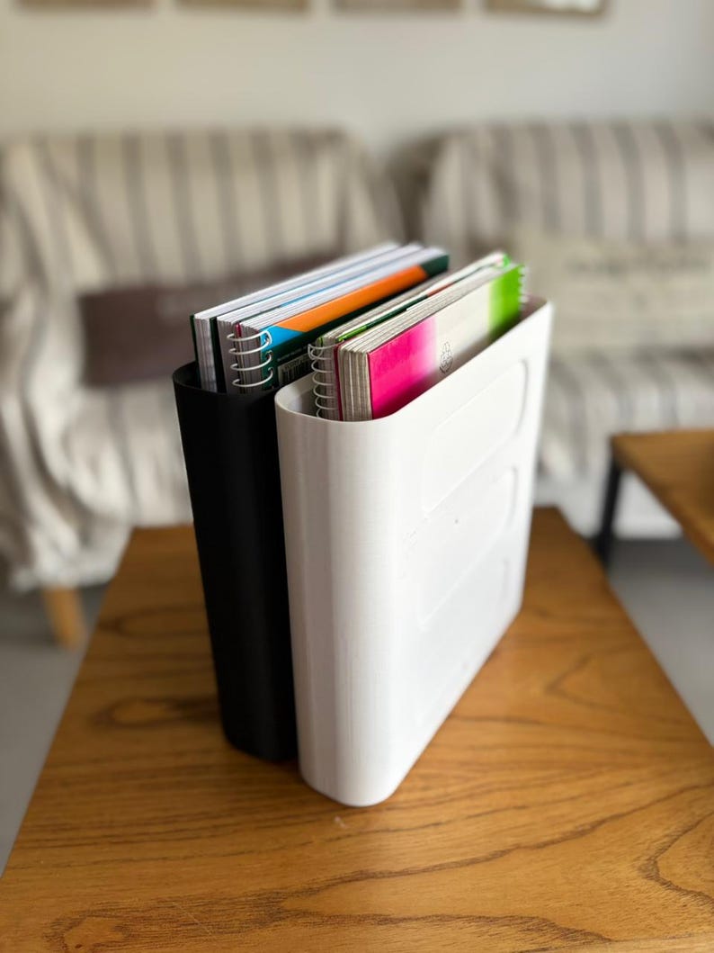 May include: Two vertical document organizers, one matte black and one glossy white, each featuring a smooth, rounded rectangular design. These organizers are filled with spiral-bound notebooks that have colorful covers in shades of orange, green, and pink.