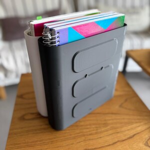 May include: A dark gray plastic vertical file organizer with three horizontal recessed rectangular panels on the front, holding several spiral-bound notebooks with colorful covers, positioned behind a light gray vertical file organizer.
