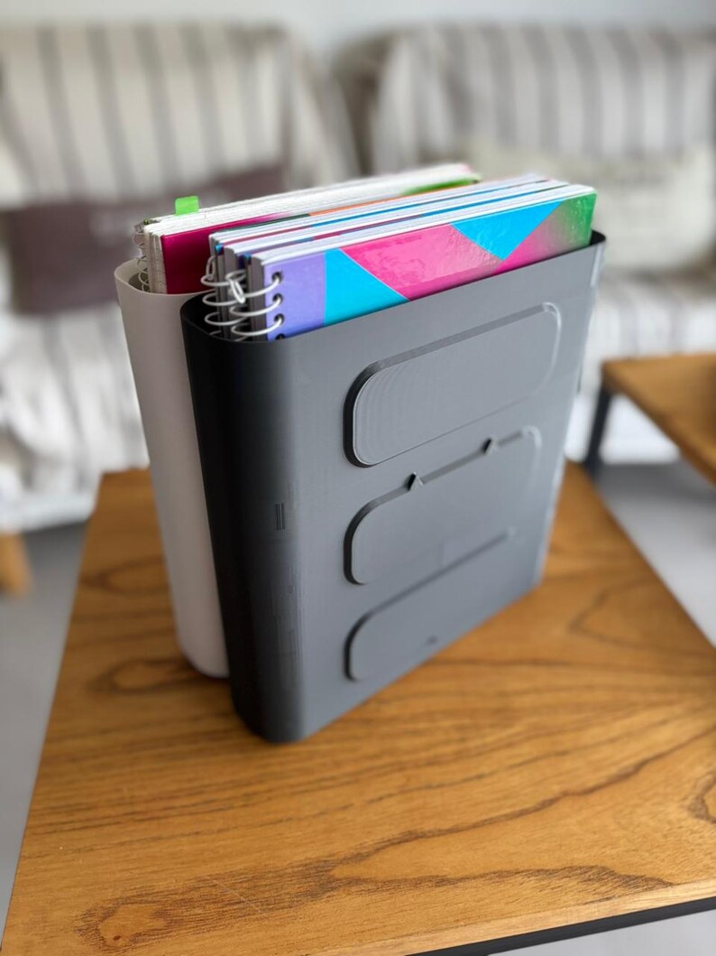 May include: A dark gray plastic vertical file organizer with three horizontal recessed rectangular panels on the front, holding several spiral-bound notebooks with colorful covers, positioned behind a light gray vertical file organizer.
